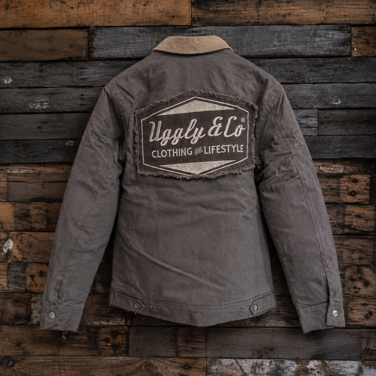 A dark gray utility jacket with a tan collar, shown from the back against a rustic wooden plank wall. The back features a large distressed patch with the brand name 'Uggly & Co Clothing and Lifestyle'.