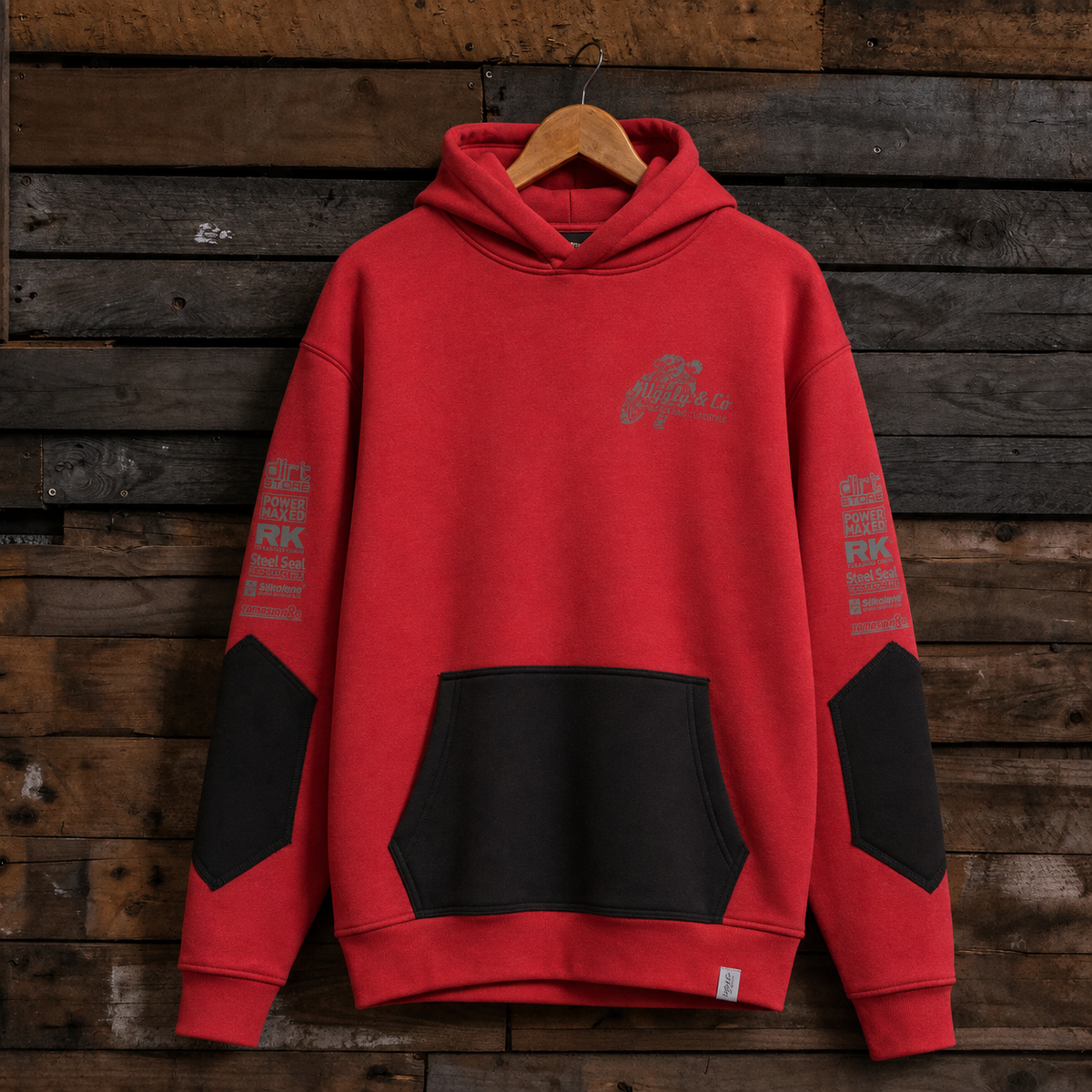 A red pullover hoodie with black front pocket and black elbow patches, hanging on a wooden hanger against a rustic dark wood plank wall. The hoodie has a small gray graphic on the left chest and gray text printed down both sleeves.
