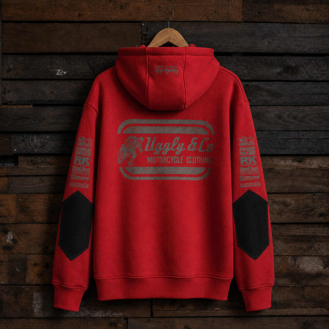 A red pullover hoodie with a large distressed gray graphic on the back reading 'Ugly & Co Motorcycle Clothing,' hanging on a wooden hanger against a dark rustic wood plank wall. The sleeves have black reinforced panels and additional distressed gray text details.