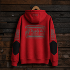 A red pullover hoodie with a large distressed gray graphic on the back reading 'Ugly & Co Motorcycle Clothing,' hanging on a wooden hanger against a dark rustic wood plank wall. The sleeves have black reinforced panels and additional distressed gray text details.