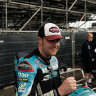 Dan Ingham in black dub hat by pit lane and race stands from side profile 