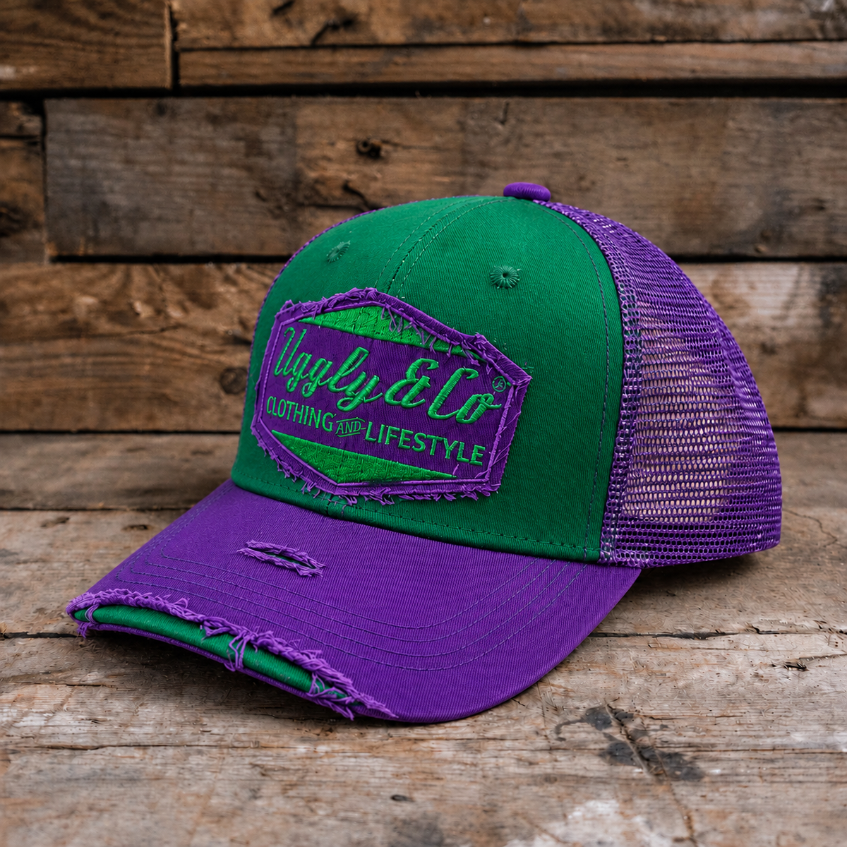 A two-tone green and purple trucker cap with a distressed brim, resting on a rustic wooden surface against a wooden plank background. The front panel features a green and purple embroidered patch reading 'Uggly & Co CLOTHING FOR LIFESTYLE'.
