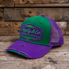 A two-tone green and purple trucker cap with a distressed brim, resting on a rustic wooden surface against a wooden plank background. The front panel features a green and purple embroidered patch reading 'Uggly & Co CLOTHING FOR LIFESTYLE'.
