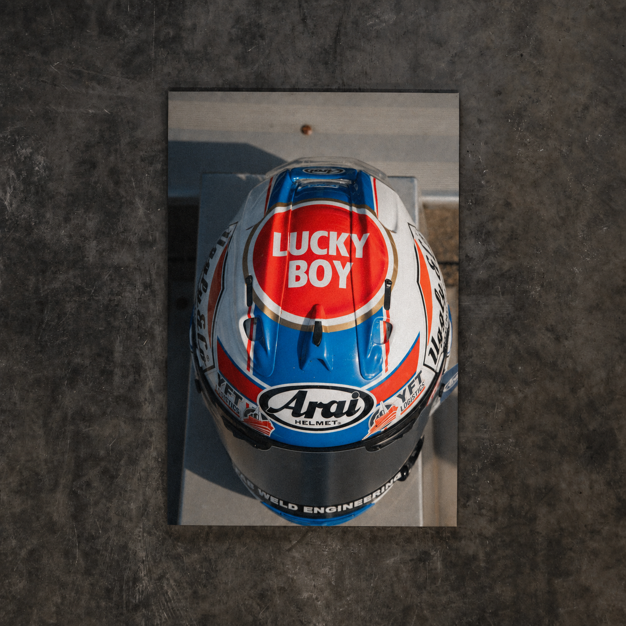 A top-down view of a racing helmet with a reflective mirror finish, featuring prominent 'Lucky Boy' and 'Arai' branding, set against a dark textured concrete background.