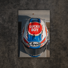 A top-down view of a racing helmet with a reflective mirror finish, featuring prominent 'Lucky Boy' and 'Arai' branding, set against a dark textured concrete background.