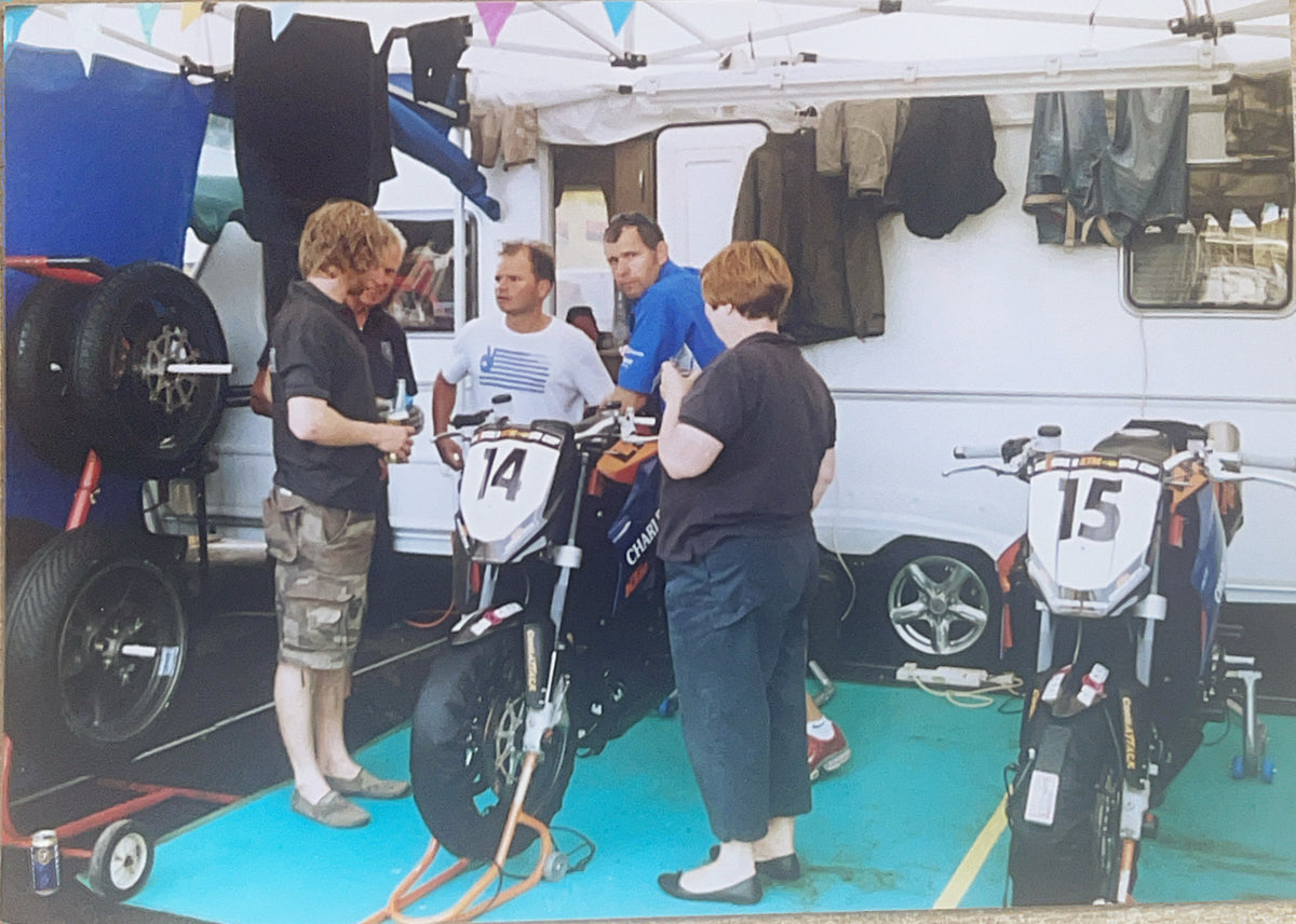 Several people gathered around two racing motorcycles at a track or event, with a white trailer and clothing hanging in the background.