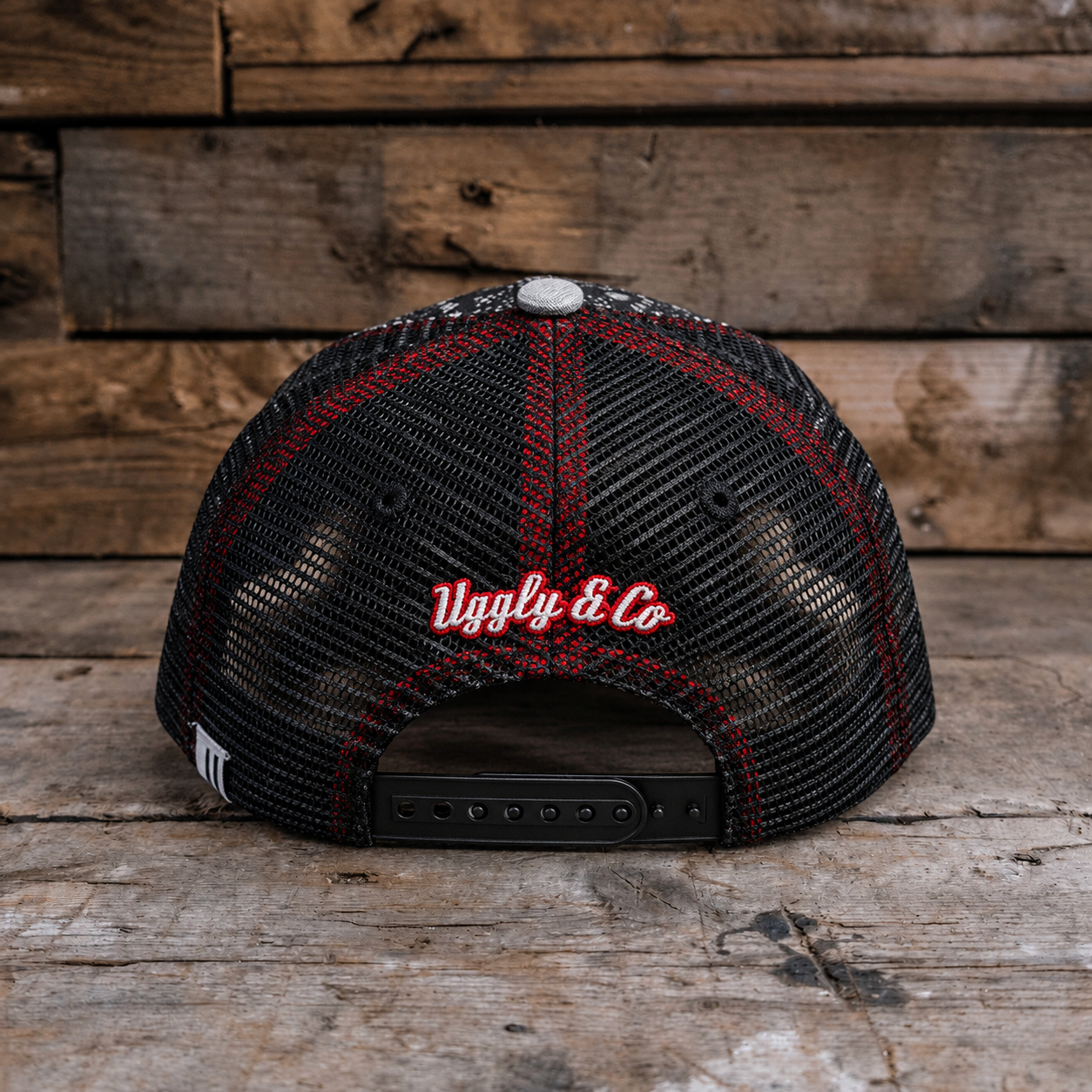 A black trucker-style cap with red mesh trim and red embroidered 'Uggly & Co' text on the back, photographed on a rustic wooden surface with a matching wood plank background.