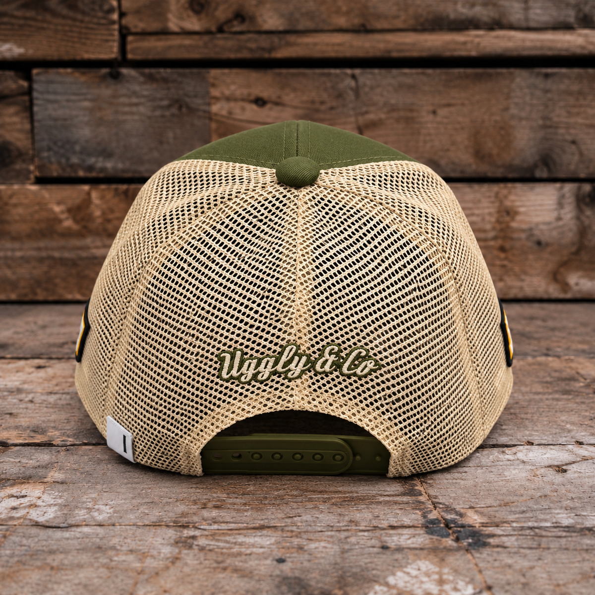 A beige mesh-back trucker cap with an olive green front panel, button, and adjustable strap, featuring 'Uggly & Co' embroidered on the back, set against a rustic wooden plank background.