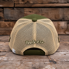 A beige mesh-back trucker cap with an olive green front panel, button, and adjustable strap, featuring 'Uggly & Co' embroidered on the back, set against a rustic wooden plank background.