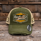 An olive green and beige trucker-style baseball cap with a distressed, frayed look, featuring a large embroidered patch on the front with a deer graphic and the text 'Uggly & Co'. The side panels have '51' printed on them, and a 'Clothing & Lifestyle' sticker is visible on the brim, set against a rustic wooden background.