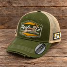 An olive green and beige trucker hat with a distressed front patch reading 'Uggly Co' and a springbok South Africa graphic, displayed on a rustic wooden surface.