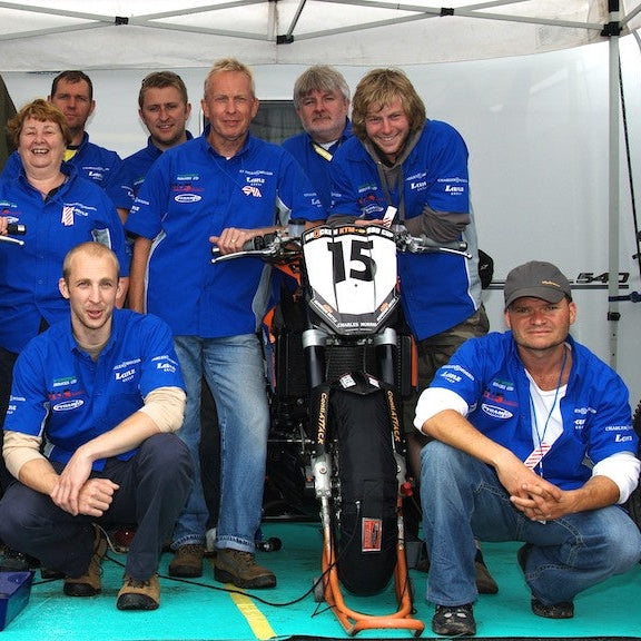 A group of people in matching blue racing team shirts posing with two off-road motorcycles numbered 14 and 15 inside a white tent.