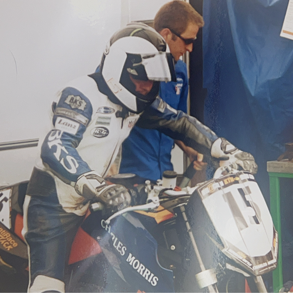 A motorcycle racer in a white, blue, and black racing suit and helmet sits on a blue and white Charles Morris motorcycle in a garage setting, with a mechanic in a blue jumpsuit standing behind him.