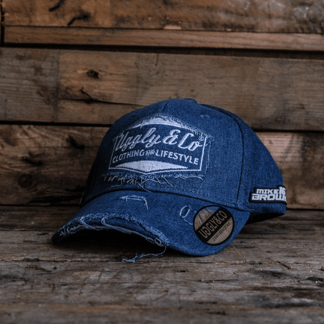 A blue denim Baseball Cap with Uggly&Co branding and Mike Browne 16 logo on the side on a wooden background