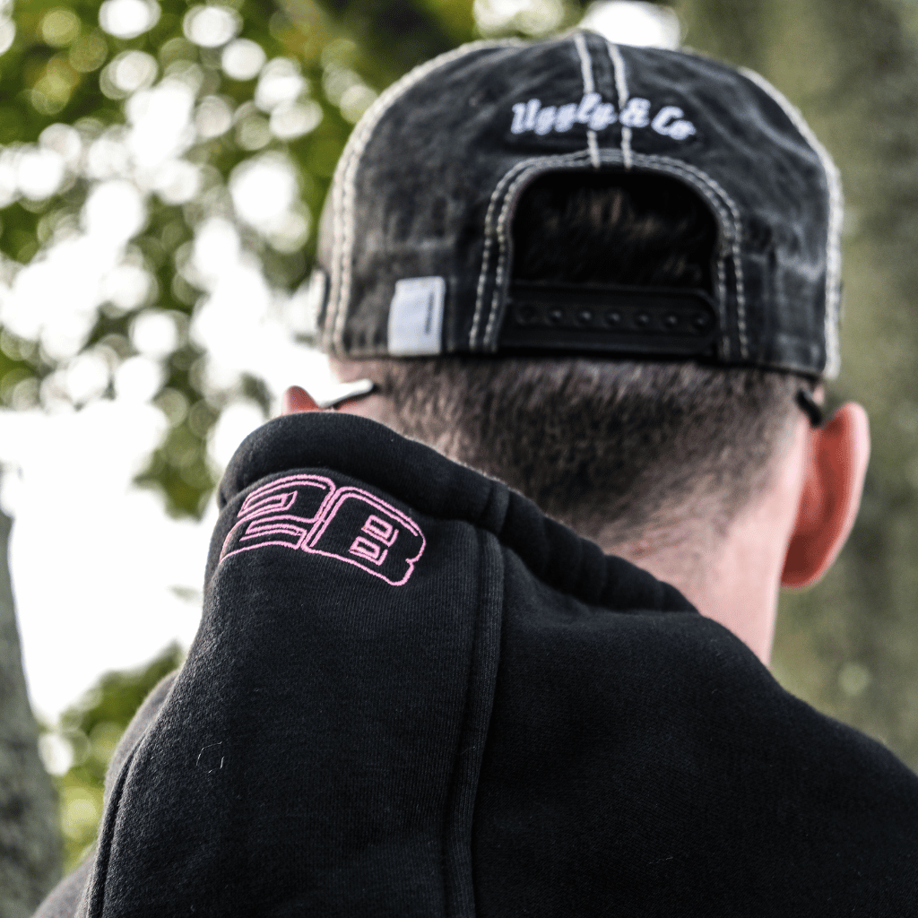 Person wearing a black cap and black hoodie with pink logo, standing outdoors.