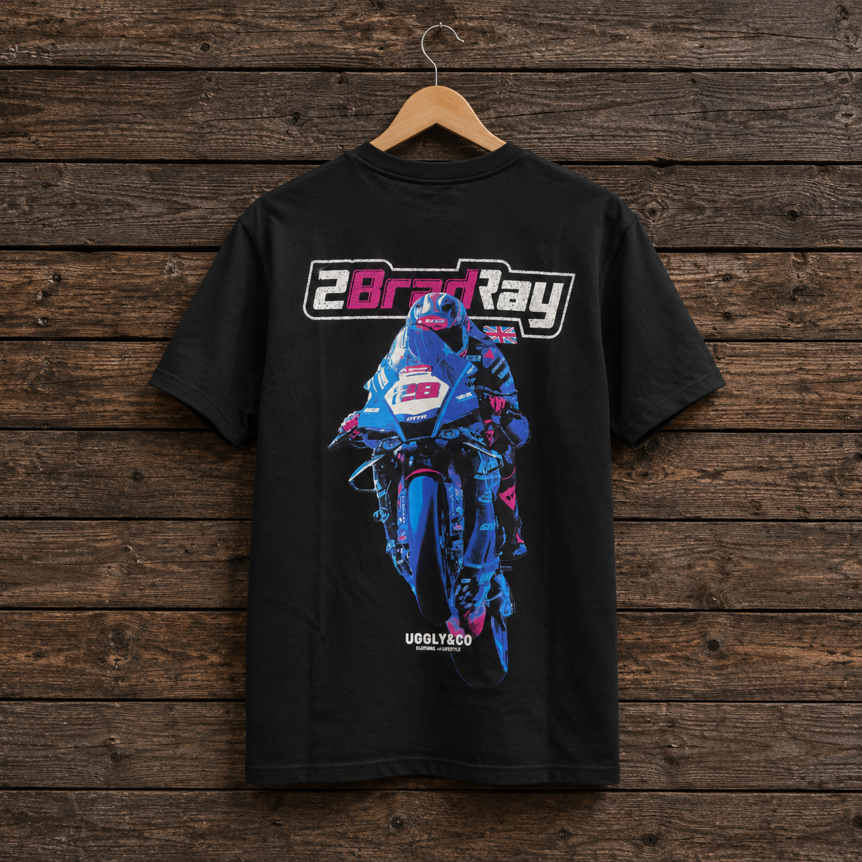 A black short-sleeve t-shirt hanging on a wooden hanger against a rustic dark wood plank wall. The back of the shirt features a large graphic print of a blue and pink motorcycle racer with the text '2BradRay' above it and 'UGGLY CO' below it.