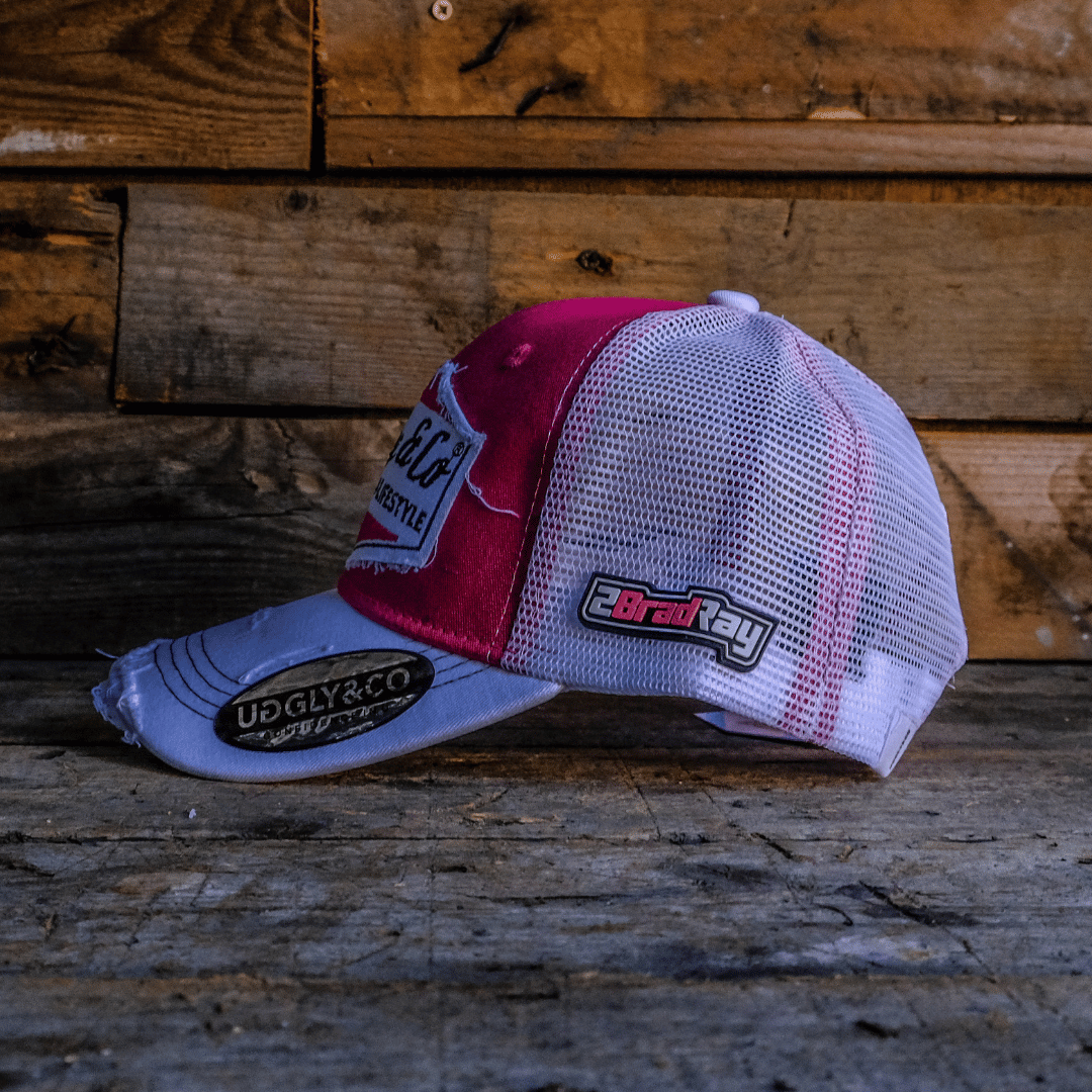Side view of a Bradley Ray branded pink and white cap with Uggly&Co diamond logo on the front on a wooden background