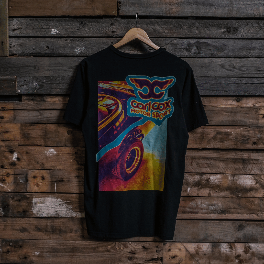 Rear view of a Carl Cox motorsport black t-shirt with a colourful graphic design on a wooden wall