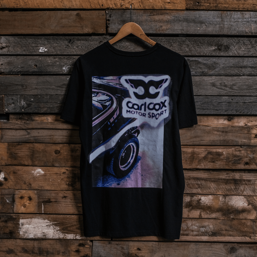 Rear view of a Carl Cox Motorsport black t-shirt with car graphic and 'carl cox motorsport' logo on a wooden background