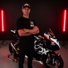 A model wearing a Carl Cox motorsport black t-shirt with an Uggly&Co logo facing the camera with a motorcycle behind