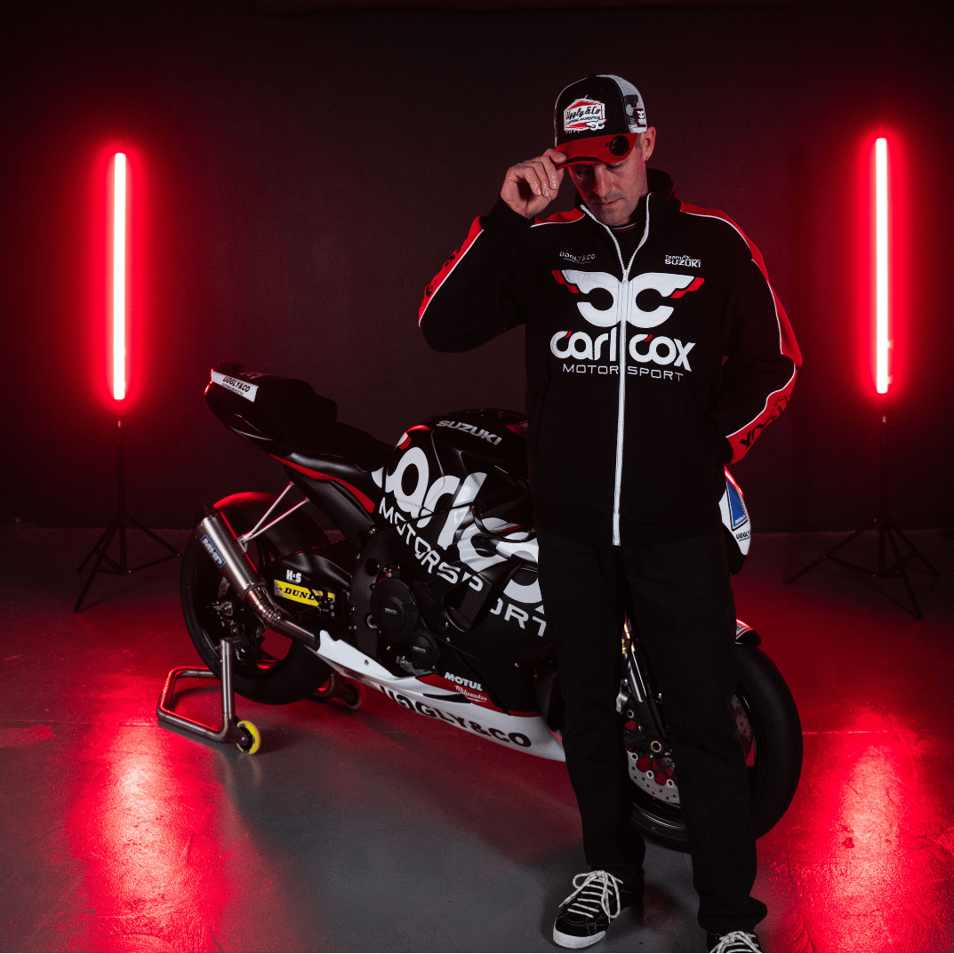Josh Brookes wearing black and red jacket with Carl Cox Motor Sport, Uggly&Co and Team Classic Suzuki branding in front of a motorbike