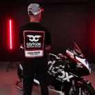 Rear view of a model wearing a Carl Cox motorsport black t-shirt with an Uggly&Co logo with a motorbike in the background