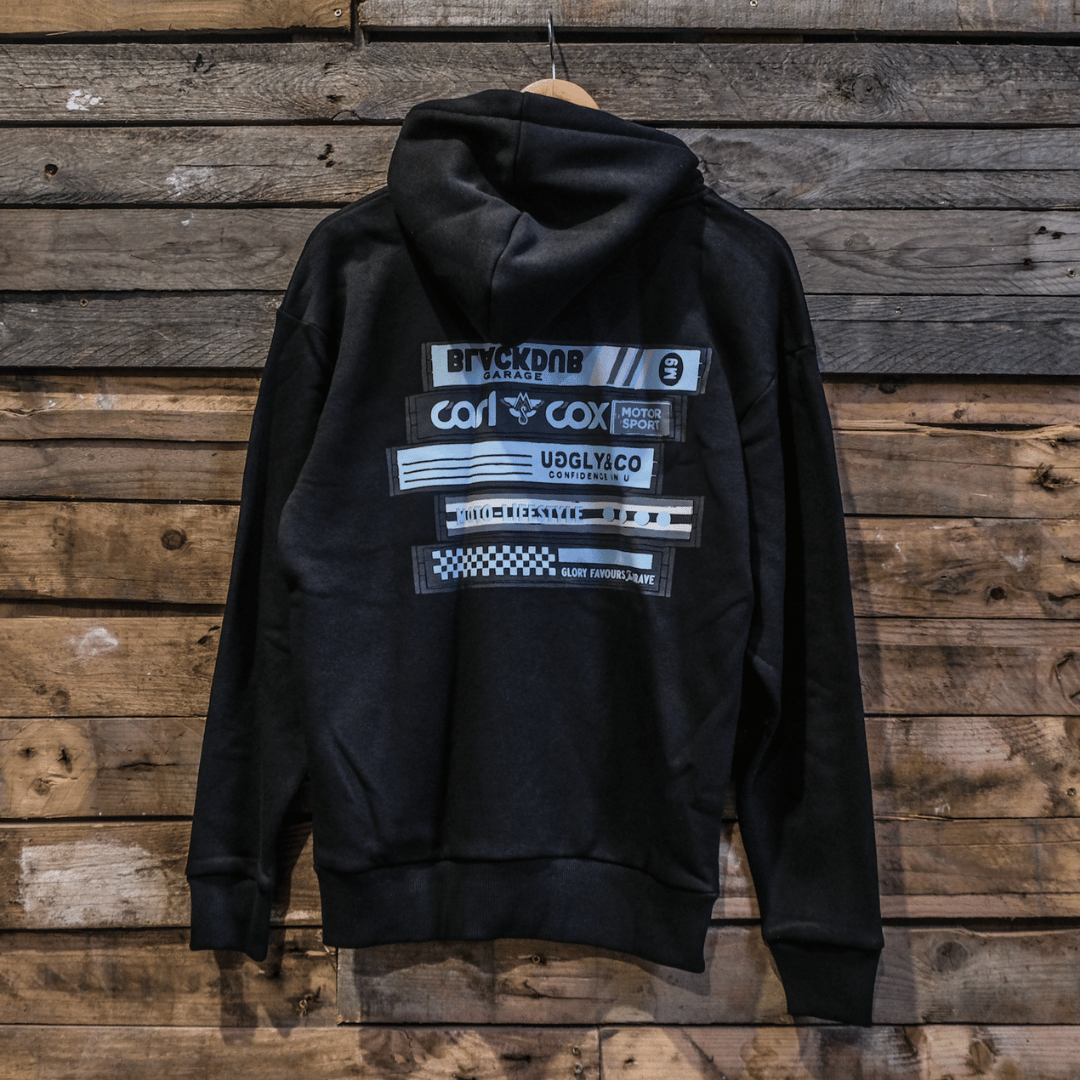 A black zip hoodie with text and graphic logos on the back, displayed against a wooden background.