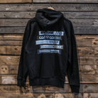 A black zip hoodie with text and graphic logos on the back, displayed against a wooden background.