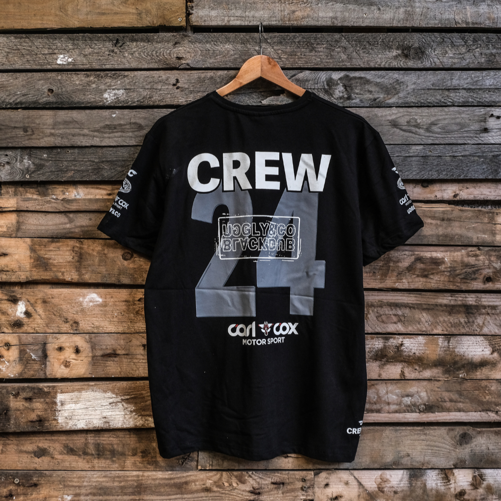 A black crew t-shirt with the text 'CREW 24' and logos printed on the front, displayed on a wooden background.