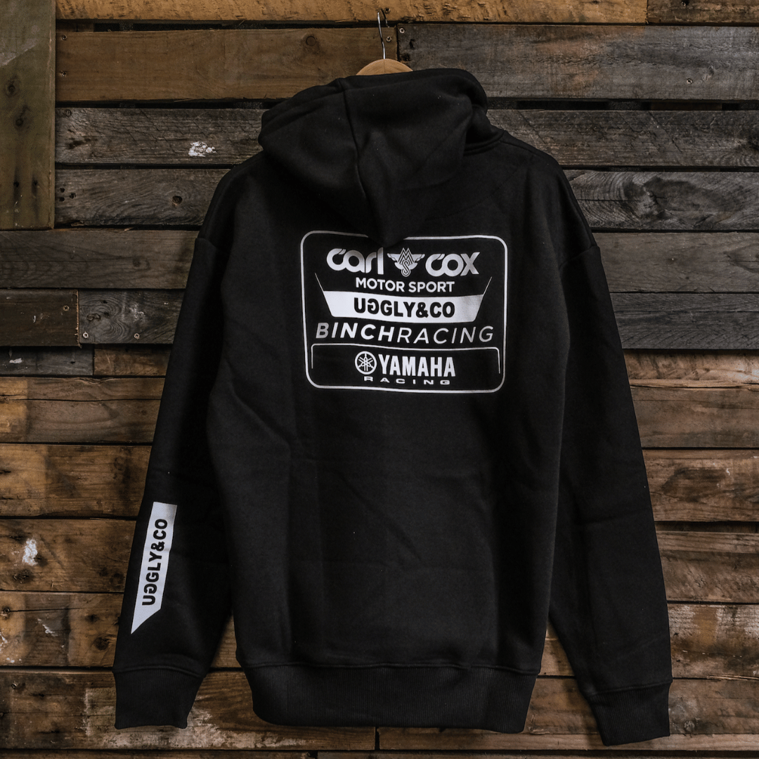 A black hoodie with white and grey text and logos, including 'Carl Cox Motorsport' and 'Uggly&Co', displayed on a wooden background.