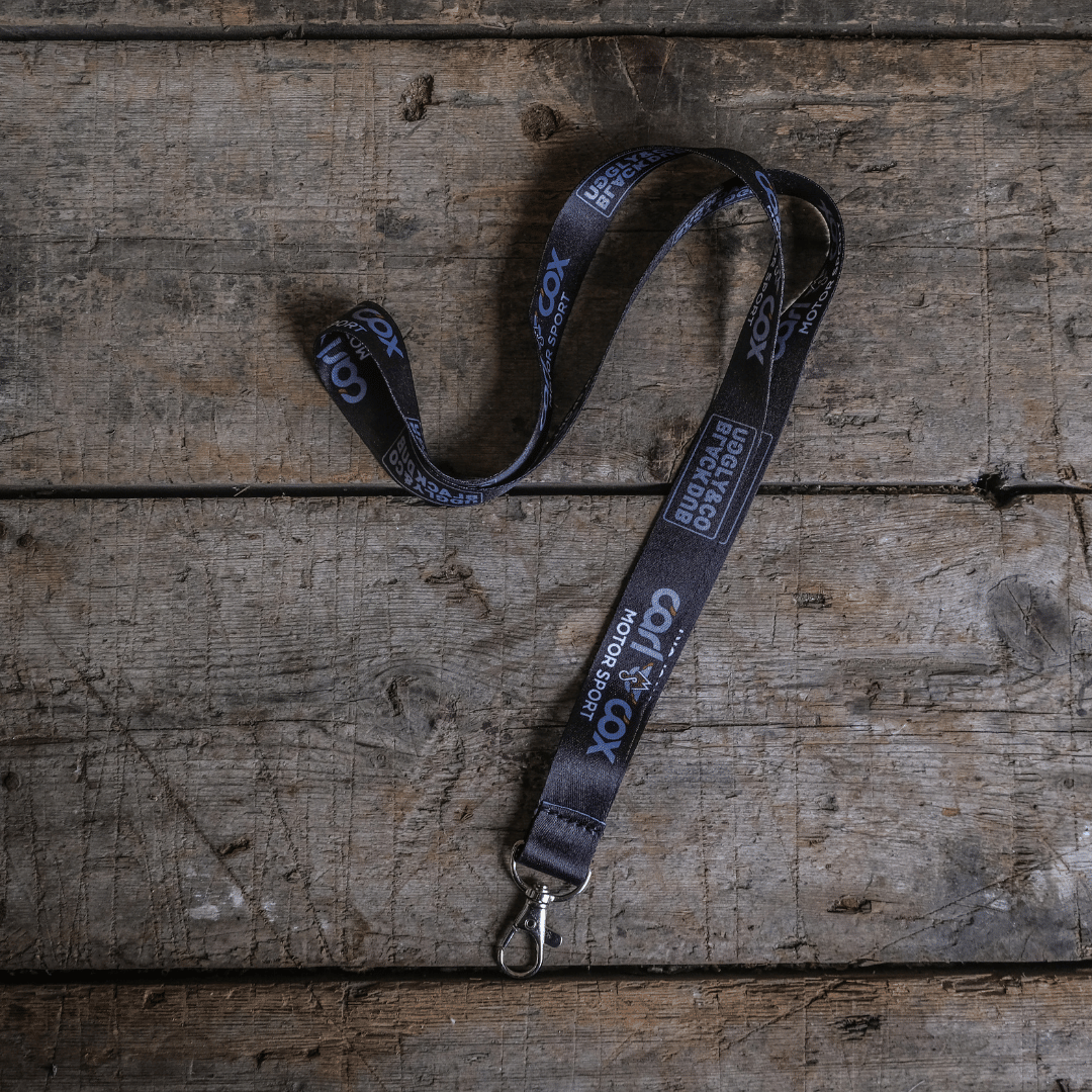 A black and silver lanyard with text and logos related to Carl Cox Motorsport and The Black Dub, laid out on a wooden surface.
