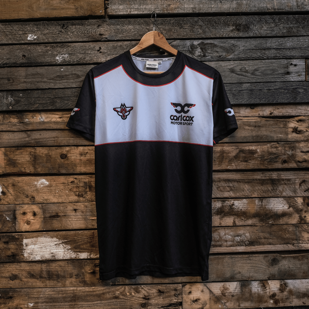A black, gray, and white t-shirt with the Carl Cox Motorsport and Uggly&Co logos displayed on the front and sleeves, hanging on a wooden backdrop.