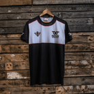 A black, gray, and white t-shirt with the Carl Cox Motorsport and Uggly&Co logos displayed on the front and sleeves, hanging on a wooden backdrop.