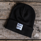 A black beanie with a white label that includes the text 'Ugglly&Co' and 'Carl Cox Motorsport' on a wooden surface.
