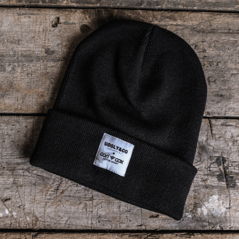 A black beanie with a white label that includes the text 'Ugglly&Co' and 'Carl Cox Motorsport' on a wooden surface.