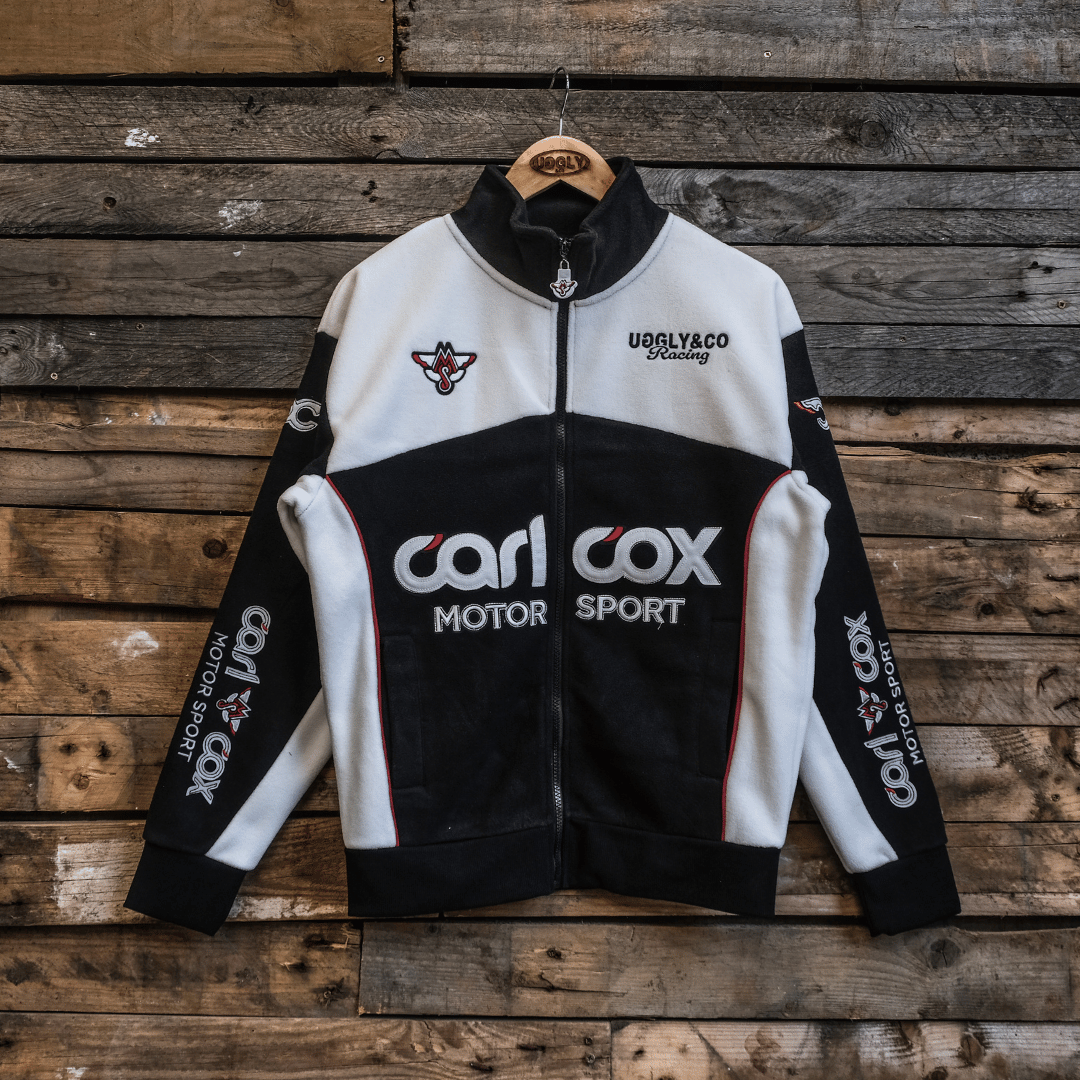 A black and white Carl Cox Motorsport track jacket with embroidered logos and badges displayed on a wooden background.