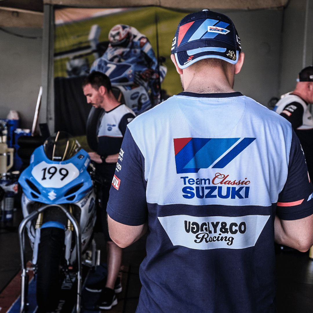A person wearing a trucker hat backwards with geometric pattern featuring blue, red, and black colours, with the Team Classic Suzuki and Suzuki logos on it in the paddock