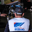 A model wearing a trucker hat backwards with geometric pattern featuring blue, red, and black colours, with the Team Classic Suzuki and Suzuki logos on it by a race track
