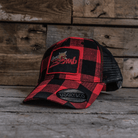 A red and black flannel trucker cap with a logo on the front, set against a wooden backdrop.