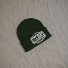 A dark forest green knit beanie with a white embroidered patch reading 'Ugly & Co Motorcycle Clothing' on a light beige fabric background.