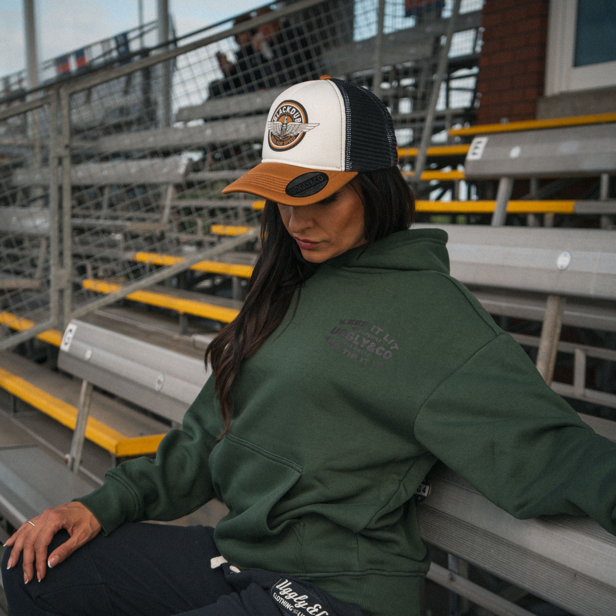 Female model wearing the green Keep It Lit hoodie in a race circuit grandstand