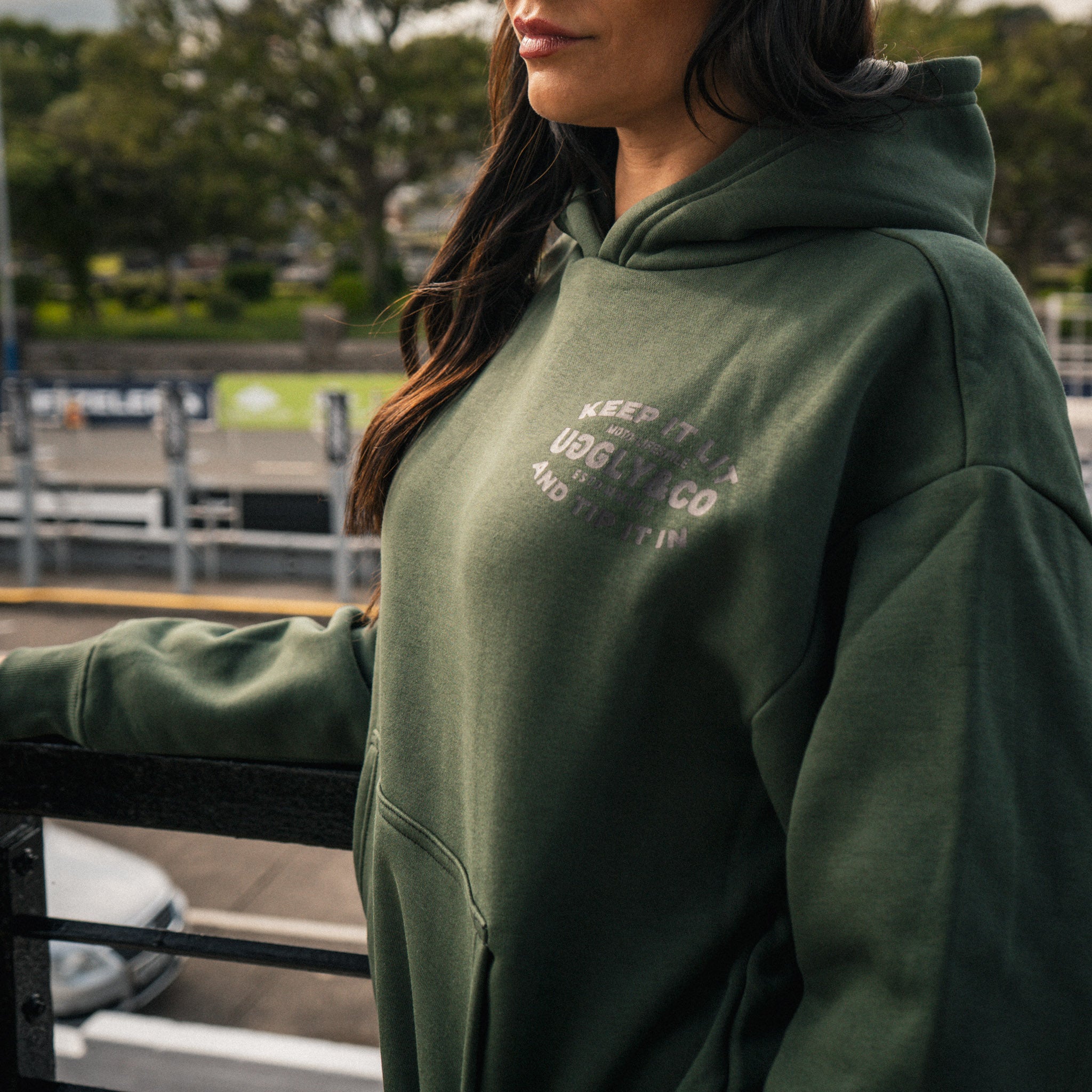 Female model wearing the green Keep It Lit hoodie beside a racetrack