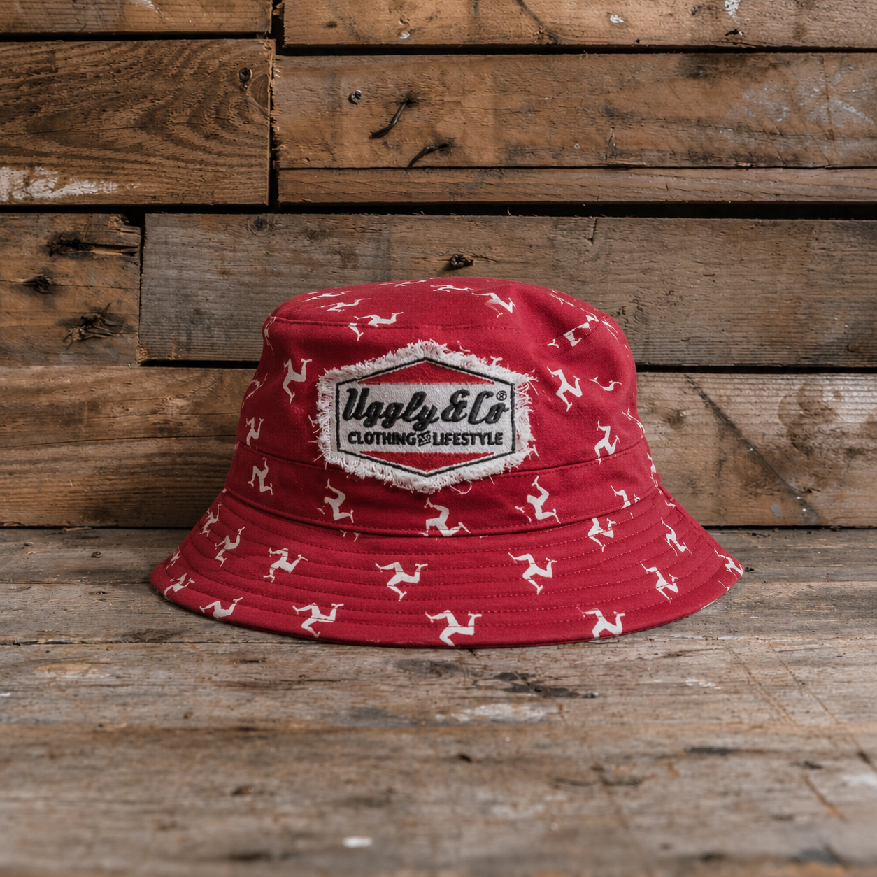 Red bucket hat with white Isle of Man Flag  patterns and a Uggly&Co logo on a wooden background