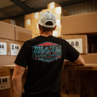 Rear view of a model wearing a Josh Brookes signature diamond black t-shirt with a blue Uggly&Co logo and a blurred background