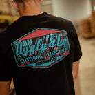 Rear view of a model wearing a Josh Brookes signature diamond black t-shirt with blue and red Uggly&Co diamond logo with a blurred background