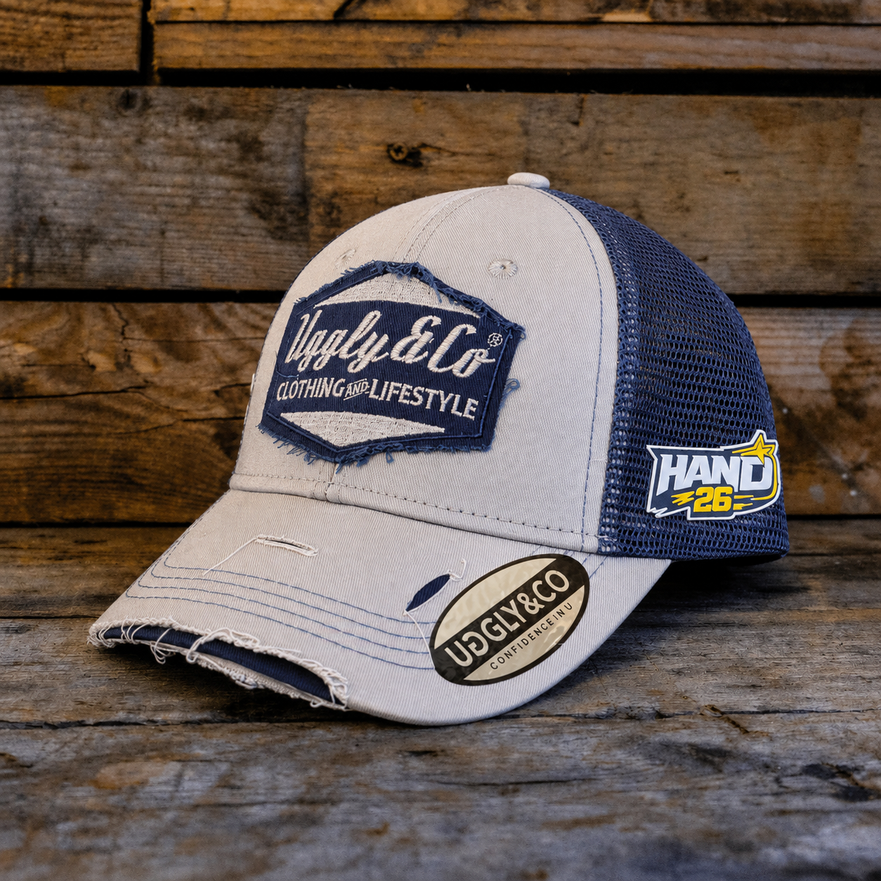 A distressed trucker cap with a light beige front and brim, navy blue mesh back panels, and embroidered patches including 'Ugly & Co' and 'HAND 26', displayed on a rustic wooden surface.