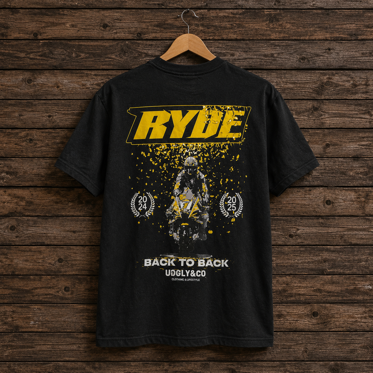 A black short-sleeve graphic T-shirt hanging on a wooden hanger against a rustic dark wood plank wall. The shirt features a large yellow 'RYDE' logo across the chest, a central motocross rider graphic with yellow splatter effects, and white text reading 'BACK TO BACK' and 'UGGLY&CO' near the hem.