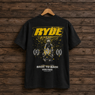 A black short-sleeve graphic T-shirt hanging on a wooden hanger against a rustic dark wood plank wall. The shirt features a large yellow 'RYDE' logo across the chest, a central motocross rider graphic with yellow splatter effects, and white text reading 'BACK TO BACK' and 'UGGLY&CO' near the hem.