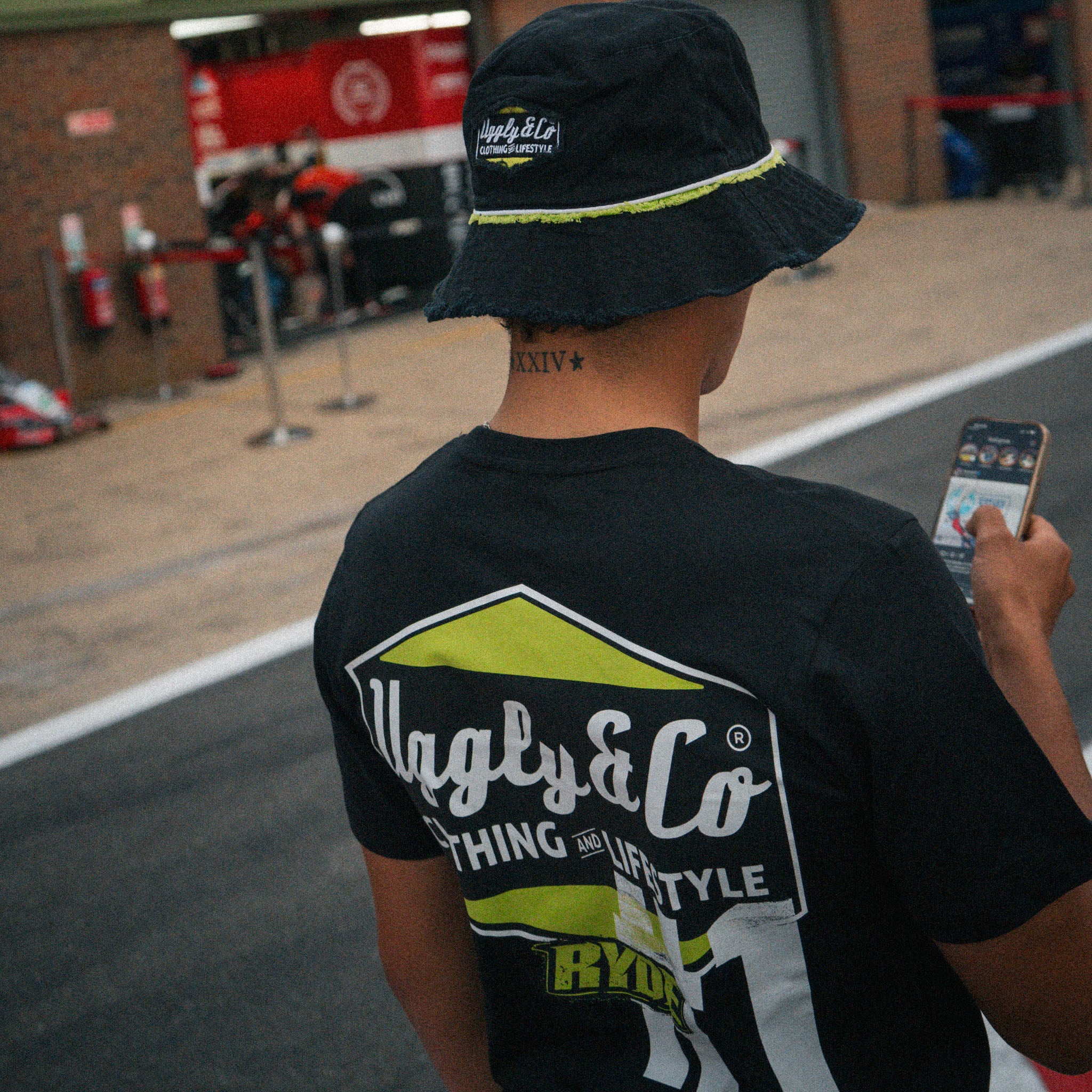 Rear view of Kyle Ryde wearing an Uggly & Co and Kyle Ryde #1 diamond graphic black t-shirt with white and lime green accents in the pit lane