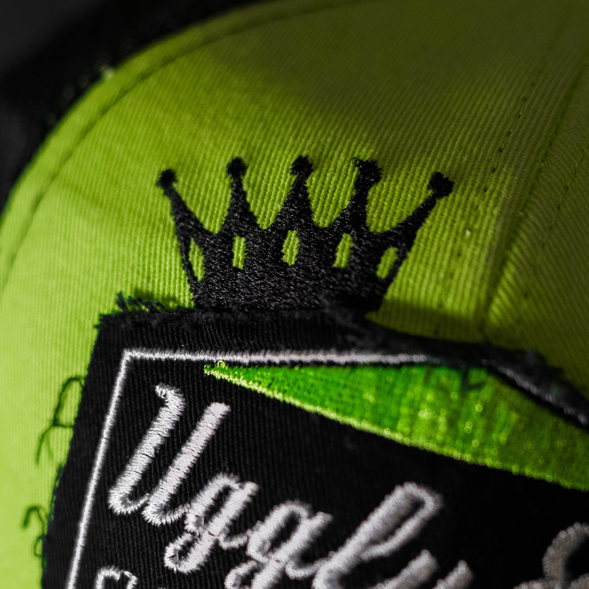 Close-up of the crown logo on the Uggly & Co Fluro Diamond Trucker Hat x Kyle Ryde, photographed against a concrete floor.